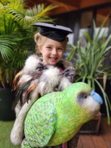 five year old child graduating preschool and on way to school wearing graduation cap and Maori feather korowai cloak next to Kakapo bird sculpture